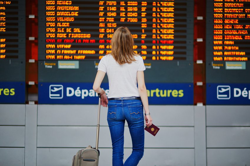 Junge Frau mit Koffer und Reisepass vor Tafel mit Abflügen