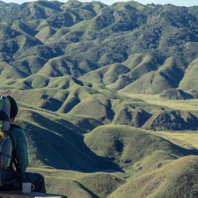 Person mit Rucksack sitzt mit einem Blick auf grüne, hügelige Landschaft