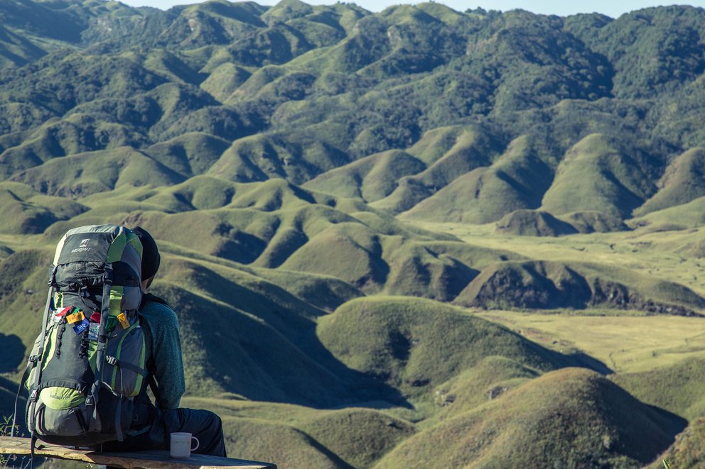 Person mit Rucksack sitzt mit einem Blick auf grüne, hügelige Landschaft